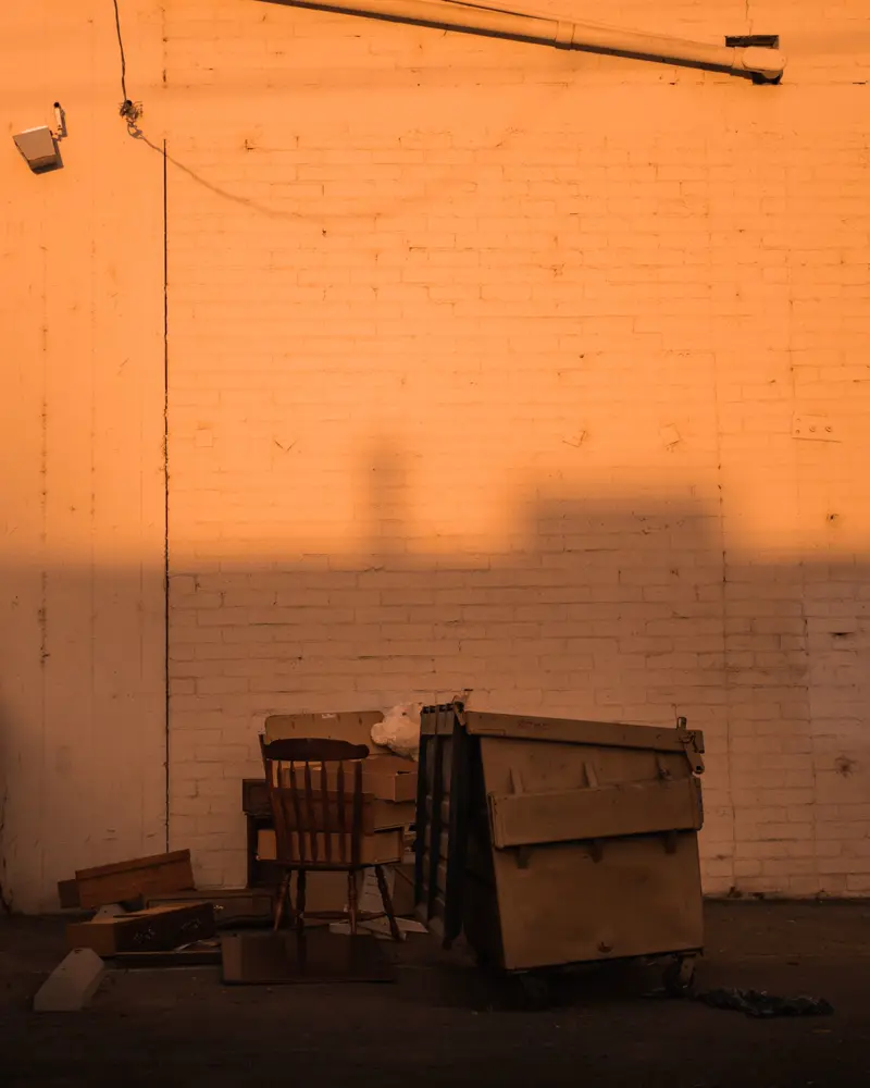 A wooden chair and discarded dresser drawers piled beside a small tan dumpster against a brick wall.