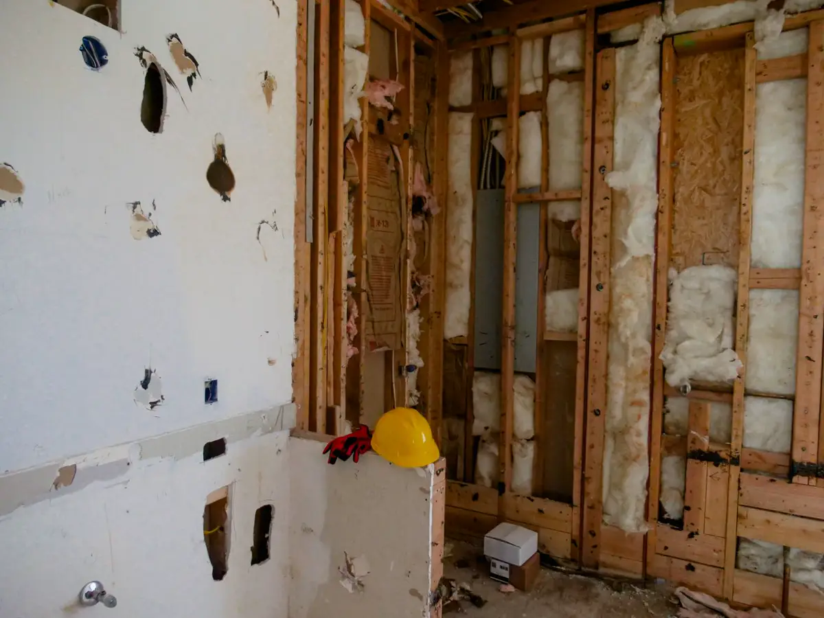 A room mid-demolition with drywall stripped to wooden studs, pink insulation, a yellow hard hat, and work gloves.